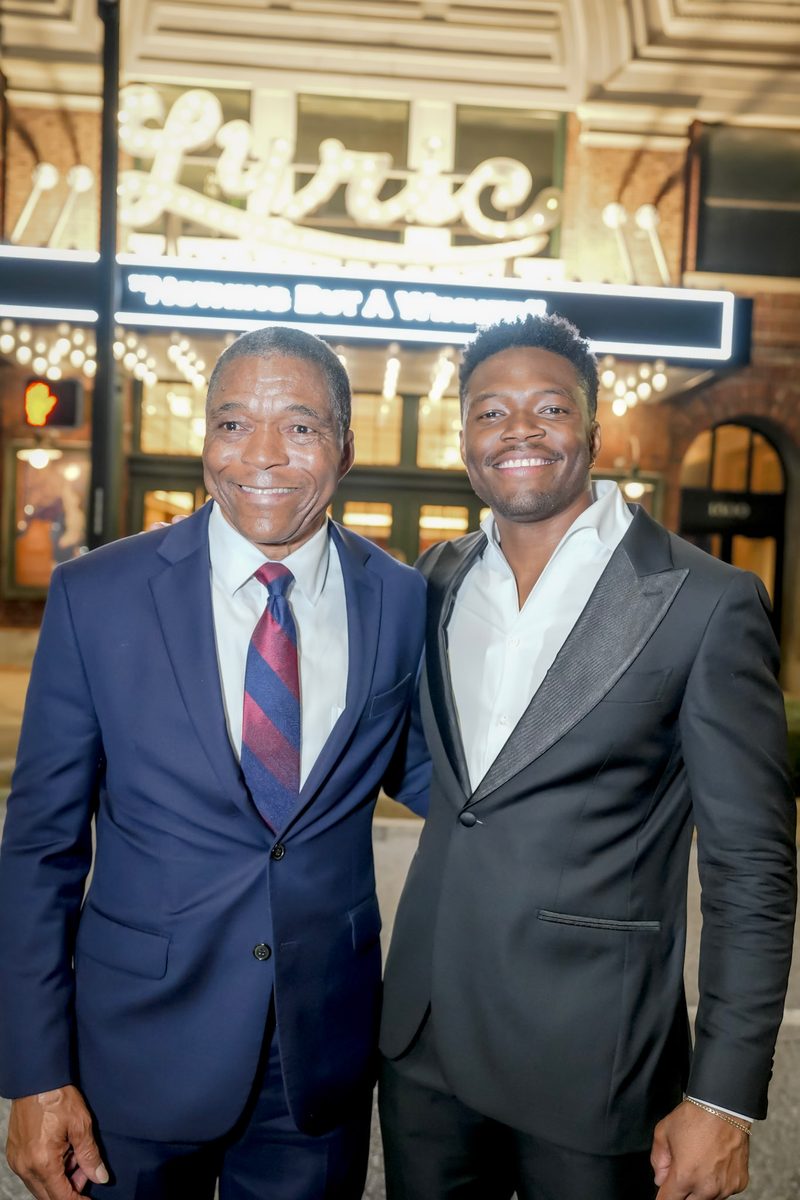 Two men in suits posing in front of a theater sign reading Lyric