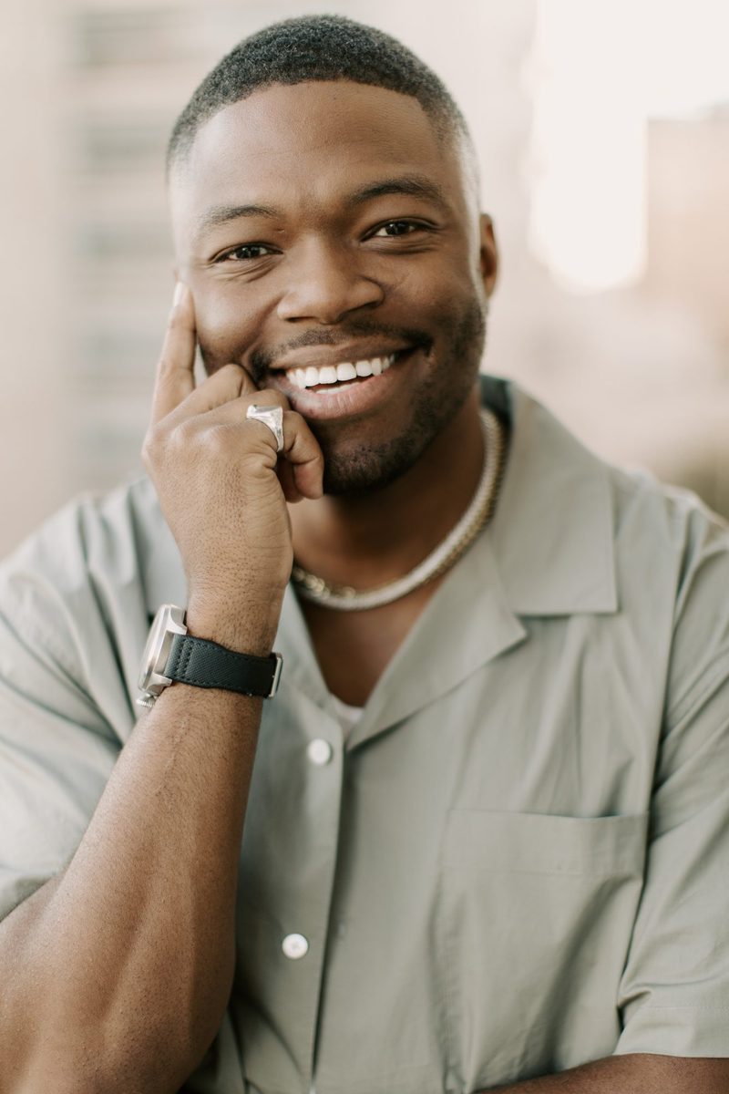 Caleb Castille smiling in an olive shirt with one hand at his face